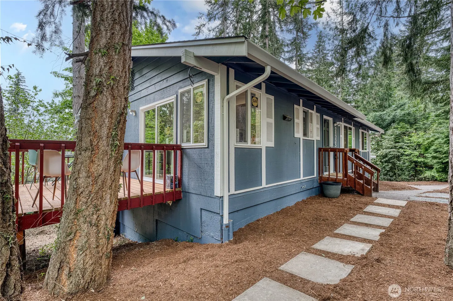 Exterior view of a blue house with a red deck and a stone walkway in a wooded area.