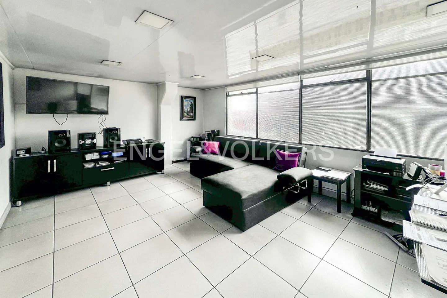Bright living room with a black sectional sofa, TV, and white tile floor. Large windows with blinds provide natural light.