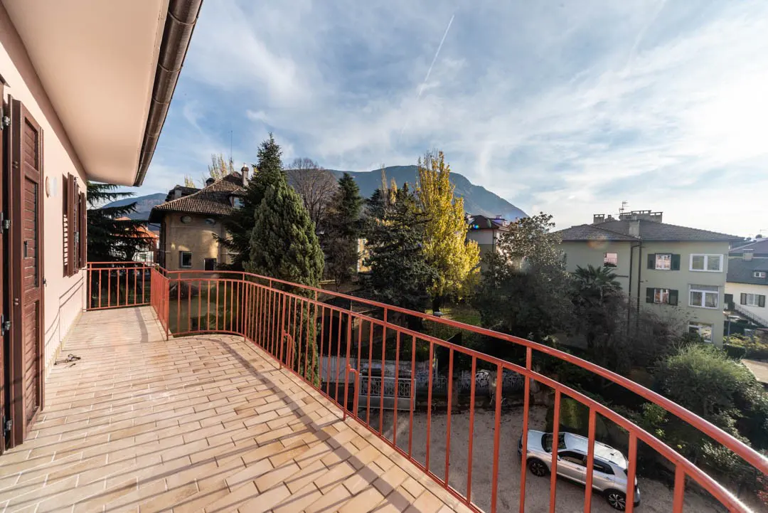 Balcony view with red railing, tan brick floor, and pink building. Trees, houses, and a mountain are in the background under a blue sky.