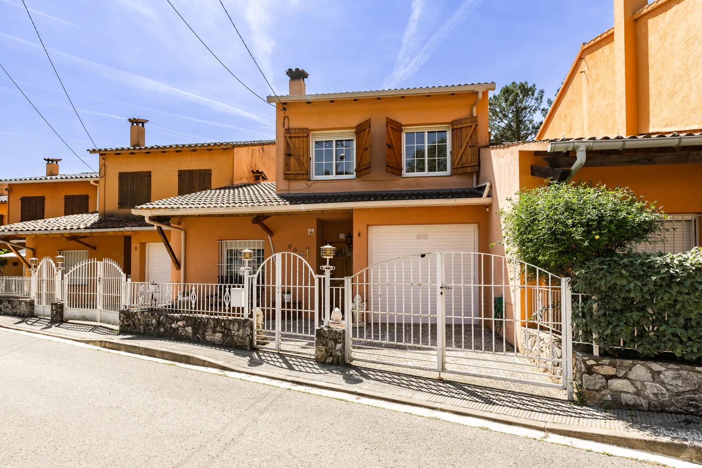 A two-story peach house with brown shutters and a white garage door, behind a white metal gate.