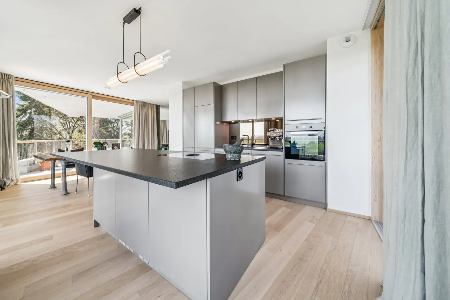A modern kitchen with gray cabinets, a black countertop island, and light wood floors. Large windows offer a view of trees and a balcony.