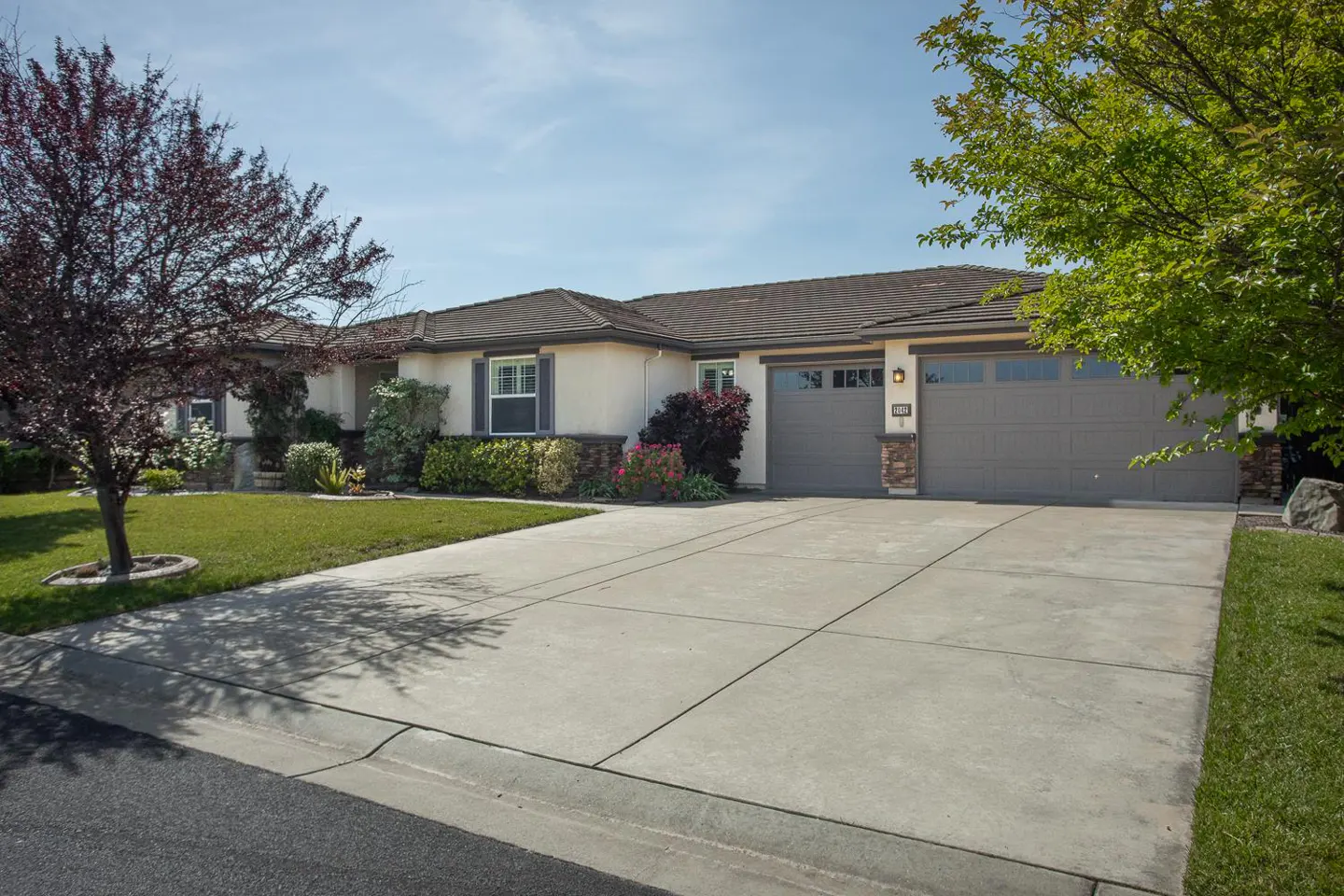 Beige single-story house with a gray two-car garage, a concrete driveway, and a green lawn on a sunny day.