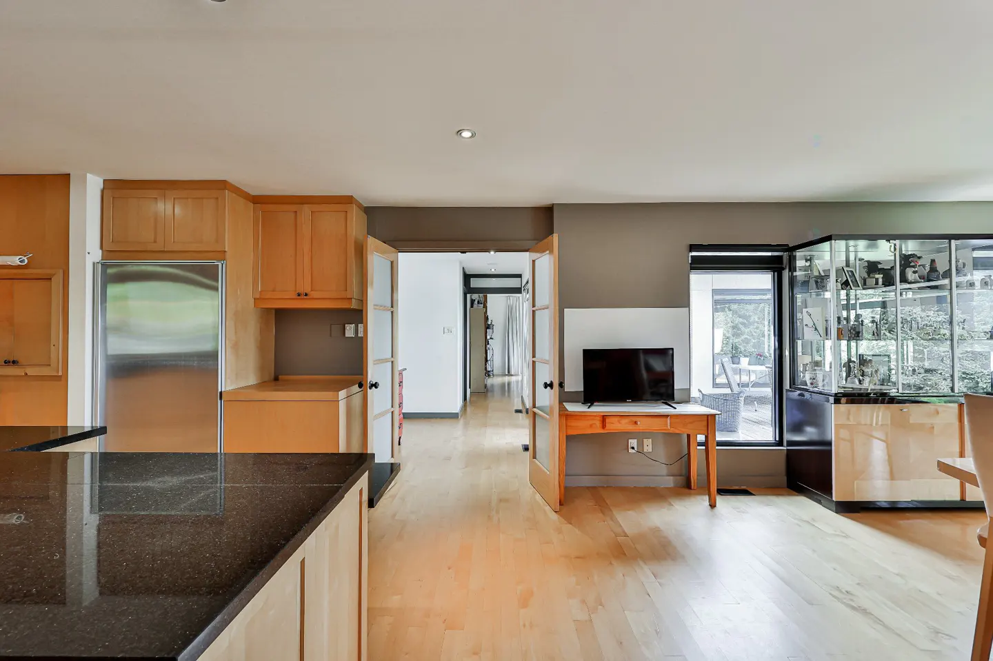 A bright, modern kitchen with light wood cabinets, stainless steel appliances, and a black granite countertop. A TV sits on a wooden table near a glass display case.