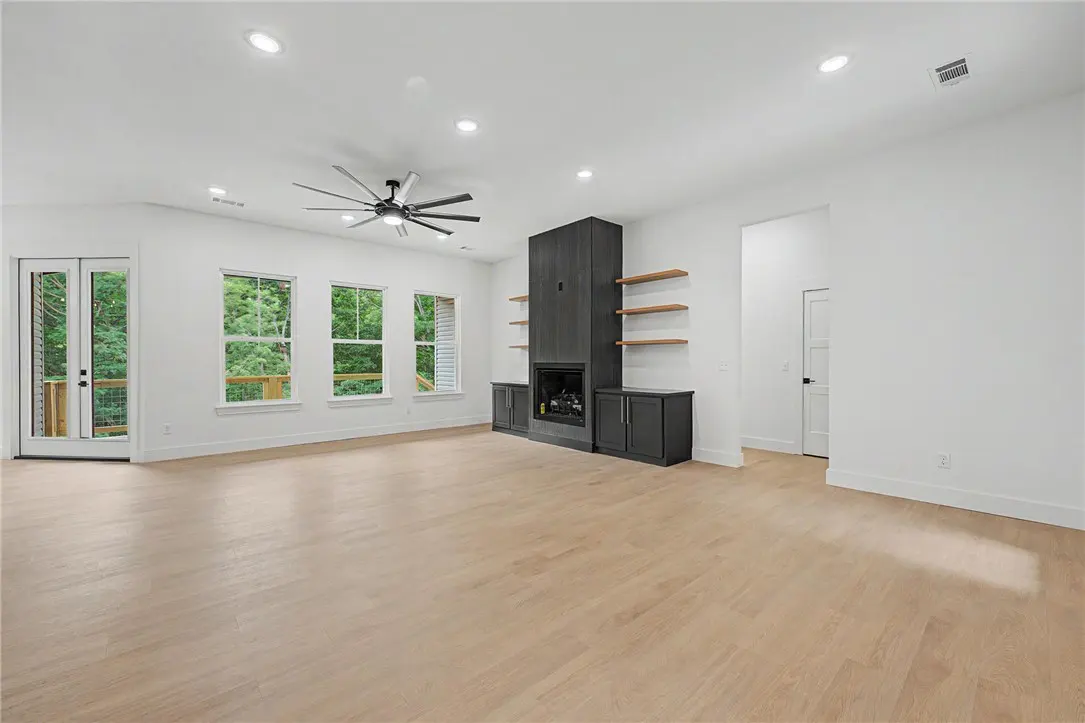 Bright, modern living room with light wood floors, white walls, and a dark gray fireplace with shelves. Windows overlook a green, wooded area.