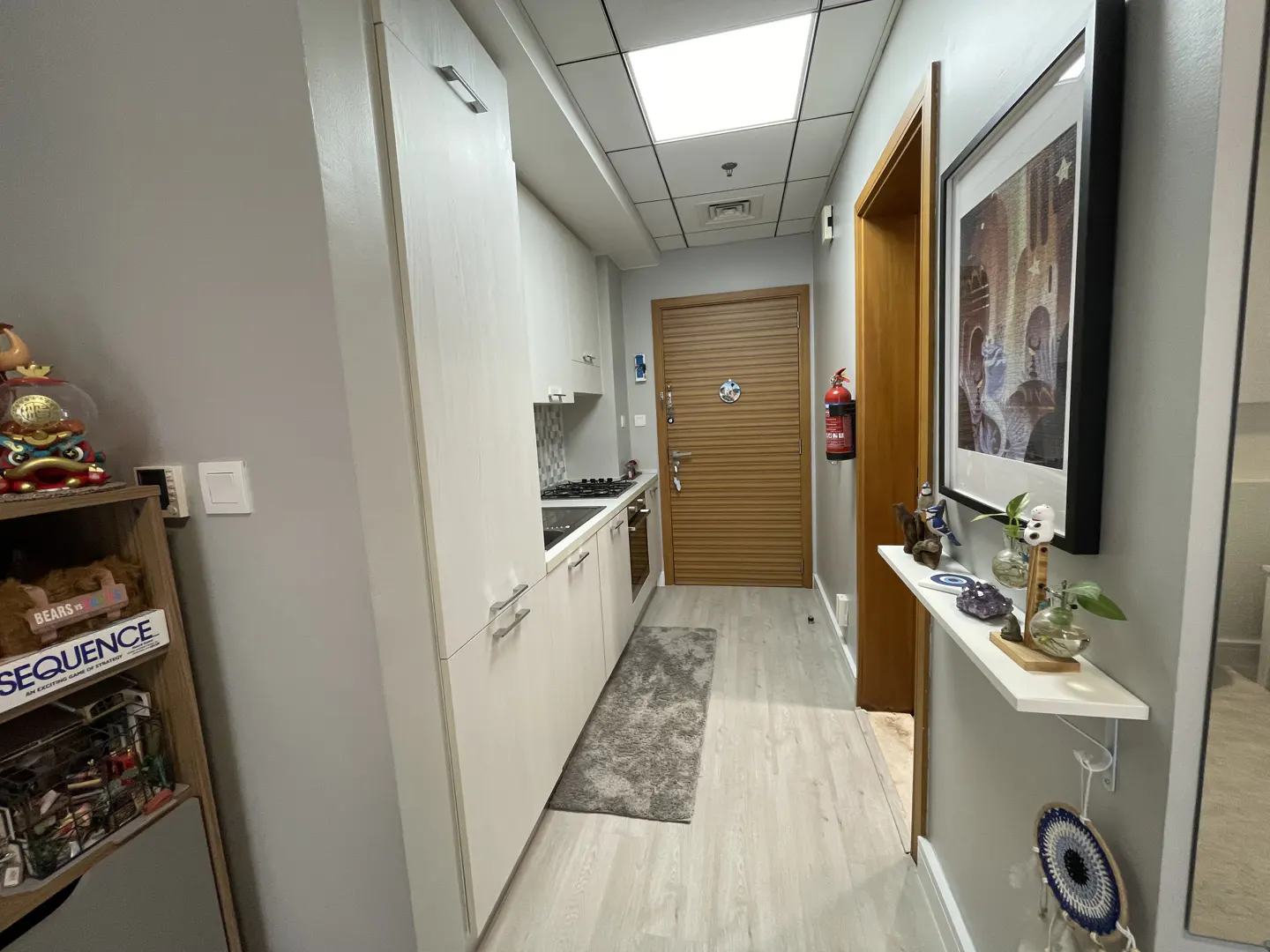 Hallway view of a modern apartment with white cabinets, light wood floors, and gray walls. A small kitchen is visible on the left.