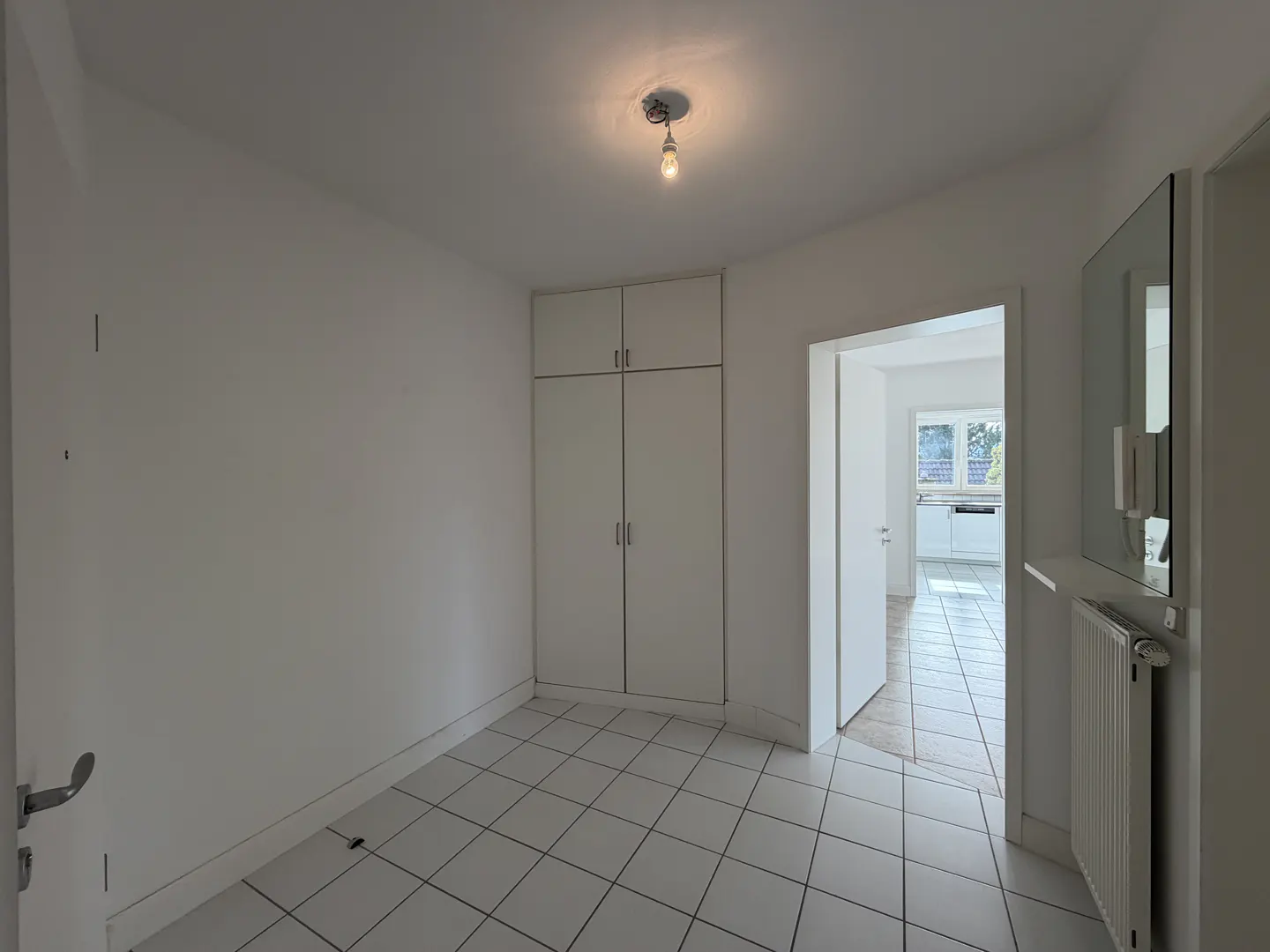 Bright, empty room with white walls, tiled floor, and a built-in wardrobe. A doorway leads to a kitchen visible in the background.