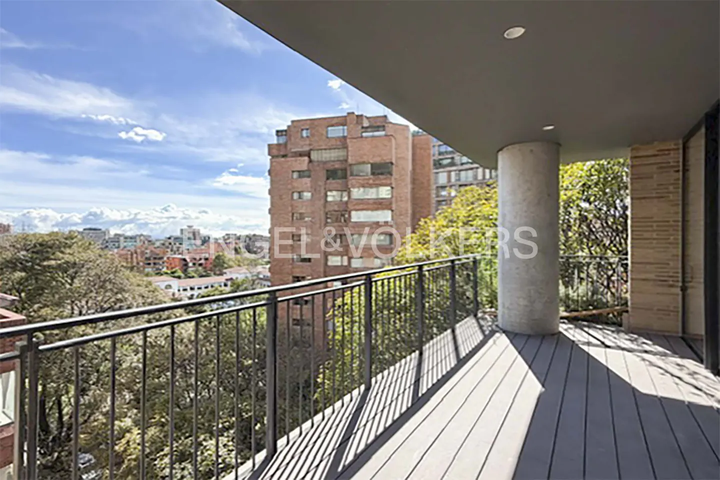 Balcony view with black railings, gray wood floor, and a concrete pillar. A brick building and city skyline are in the background.