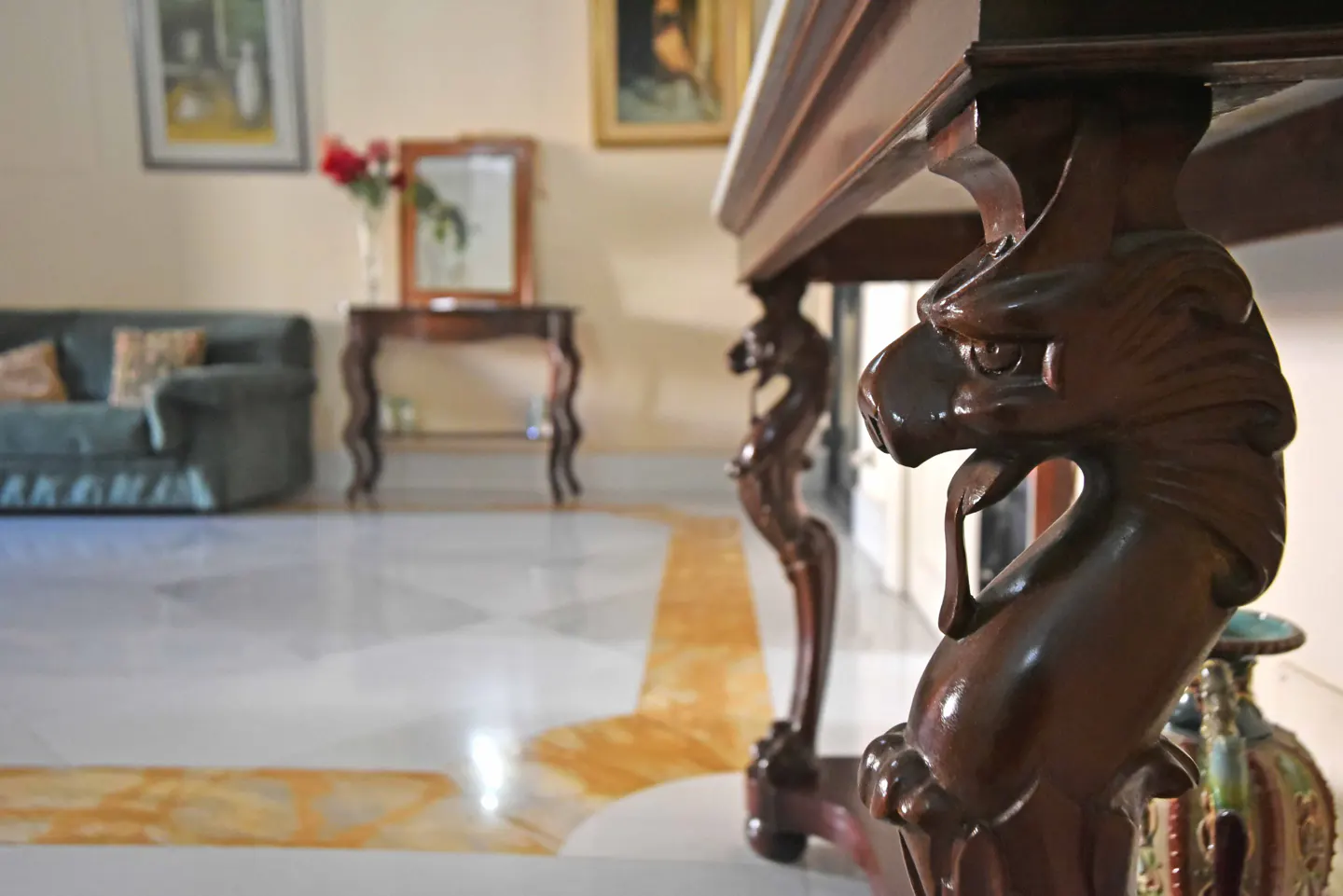 Close-up of a dark wood table leg carved with a lion's head, in a room with marble floors, a couch, and artwork.