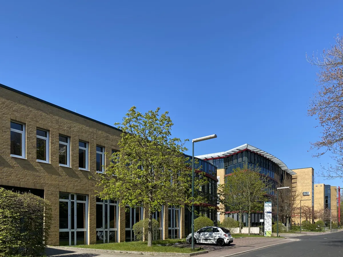 Exterior view of modern office buildings with yellow brick and glass facade under a clear blue sky. A white car is parked nearby.