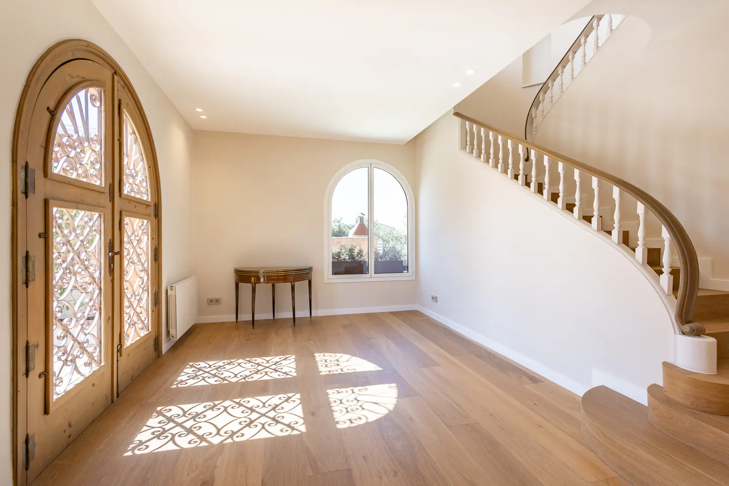 Bright foyer with wood floors, arched door with ironwork, arched window, small table, and curved staircase.