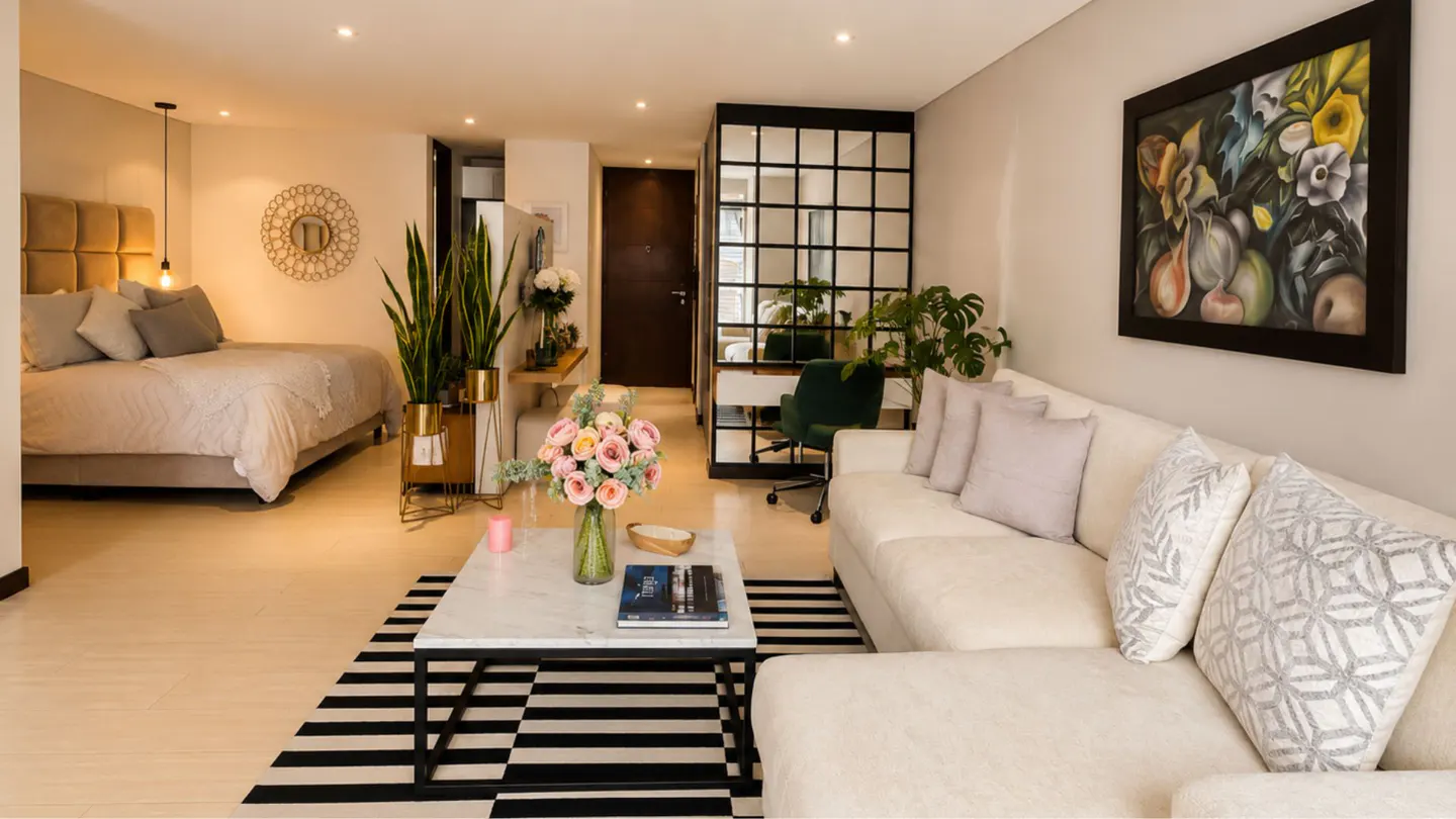 Modern studio apartment with a white sofa, marble coffee table, striped rug, and a bedroom area with a queen bed.