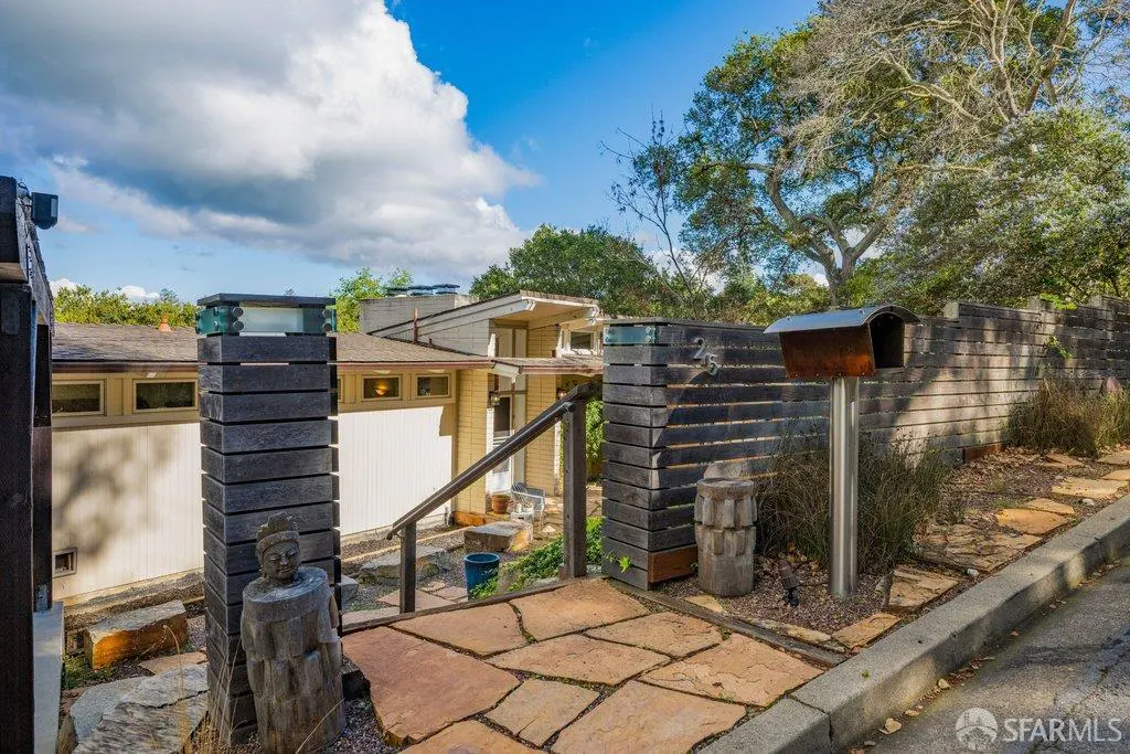 Exterior view of a house entrance with stone walkway, wooden fence, mailbox, and a statue. Blue sky with clouds in the background.