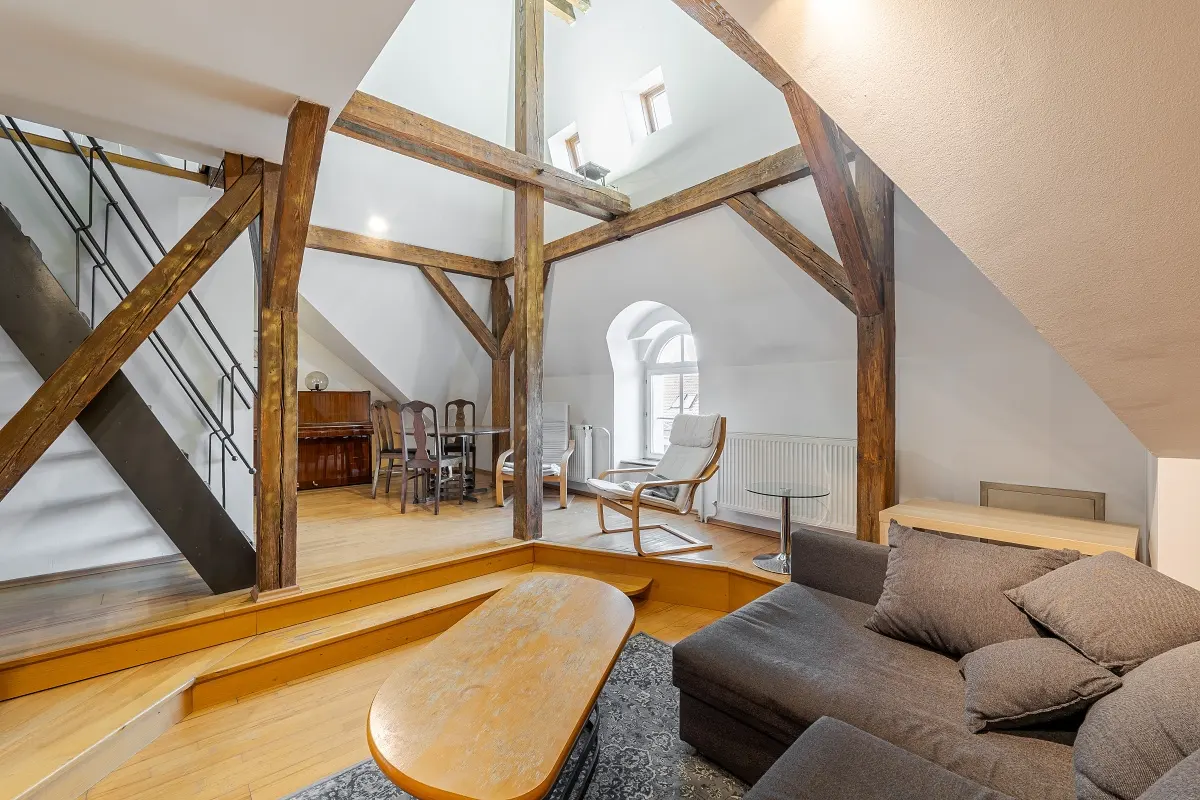 Attic living room with exposed wooden beams, gray sofa, and wooden coffee table. Stairs lead to a dining area with a piano.