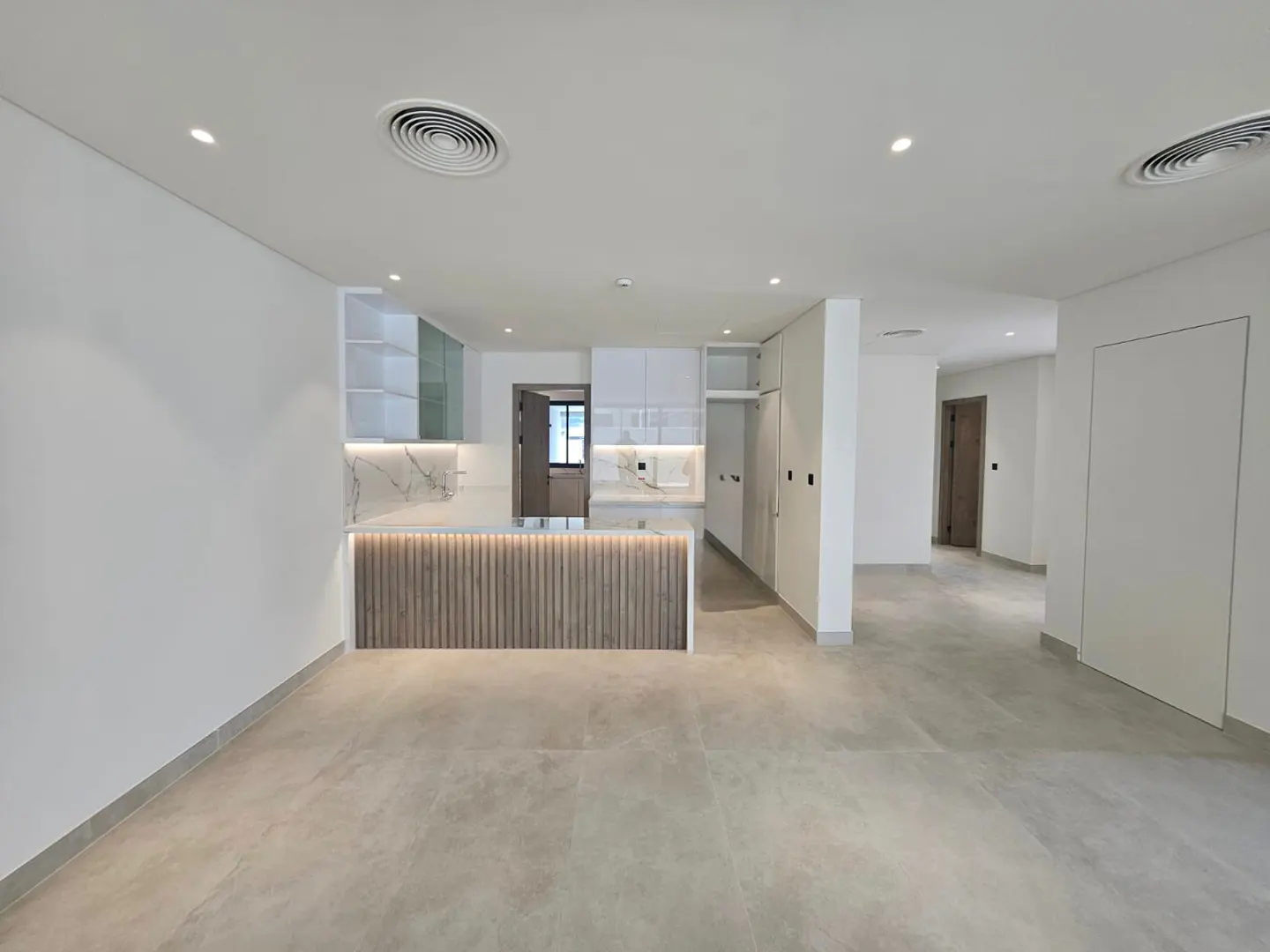 A wide shot of a modern, empty apartment with white walls, gray floors, and a kitchen with a wood-paneled island.