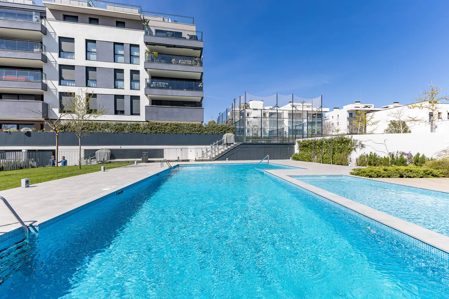 Outdoor pool with turquoise water, gray tiled deck, and modern apartment building in the background under a clear blue sky.
