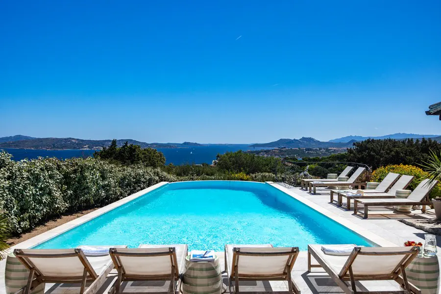 A rectangular pool with blue water, surrounded by lounge chairs, overlooks the ocean and mountains under a clear blue sky.