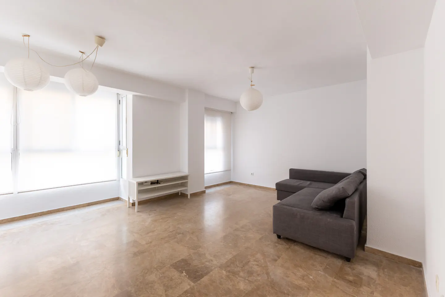 Bright, empty living room with marble floors, white walls, and a gray sectional sofa. A white shelving unit sits near a large window.