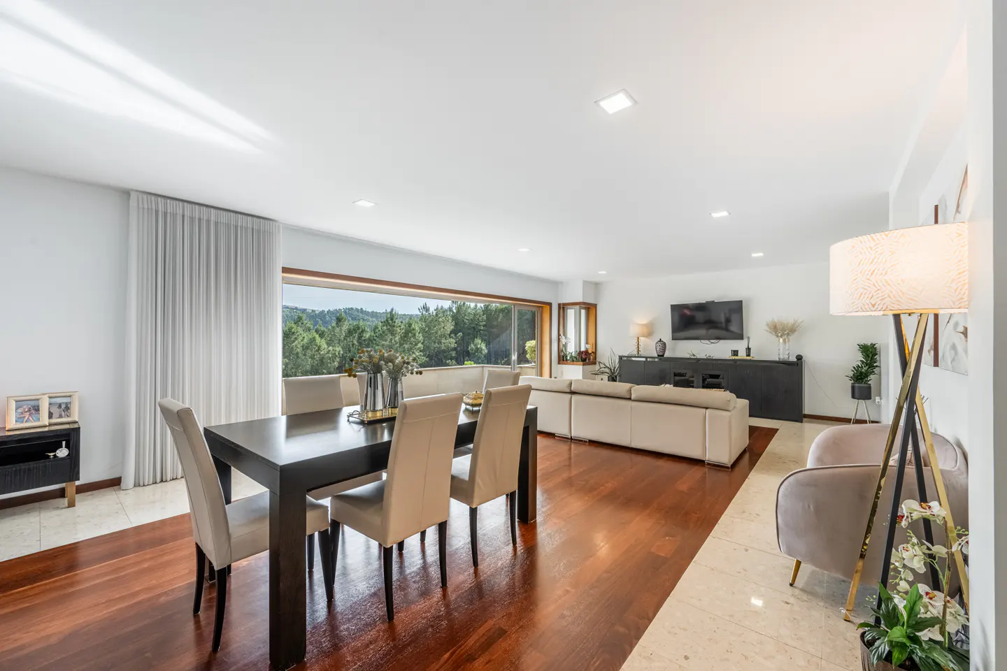 Bright, open-concept living and dining area with wood floors, a large window with a forest view, and neutral-toned furniture.