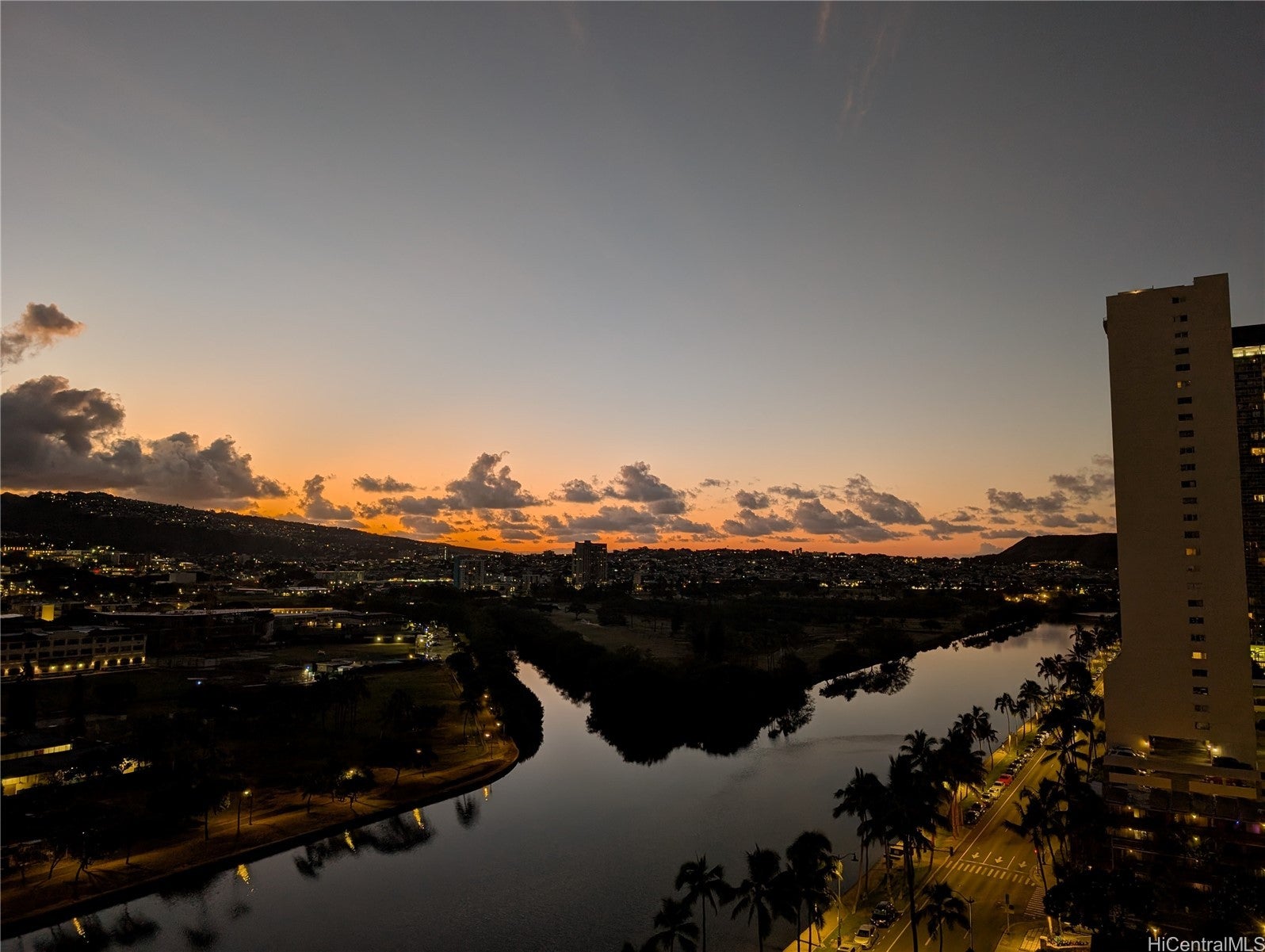 High-angle view of a city canal at sunset with a tall building on the right, palm trees, and a glowing orange horizon. [AI-generated text]