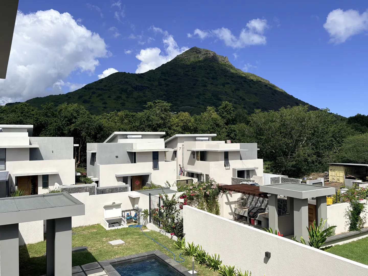 Modern white villas with gray accents, surrounded by green trees and a mountain backdrop under a blue sky with white clouds.