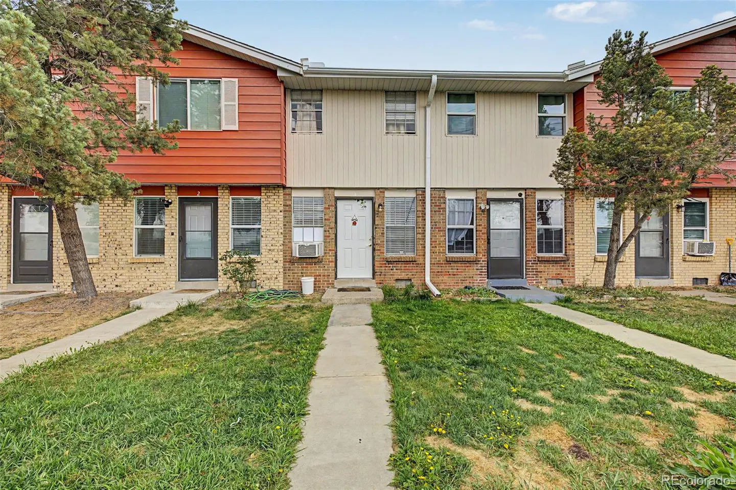 Exterior view of a row of two-story townhouses with brick and siding, a concrete walkway, and a grassy lawn.