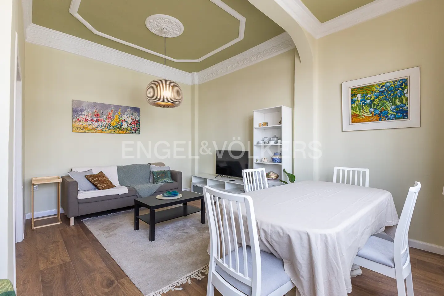 Living room with a gray sofa, black coffee table, and a dining table with white chairs. Art on the walls and a decorative ceiling.