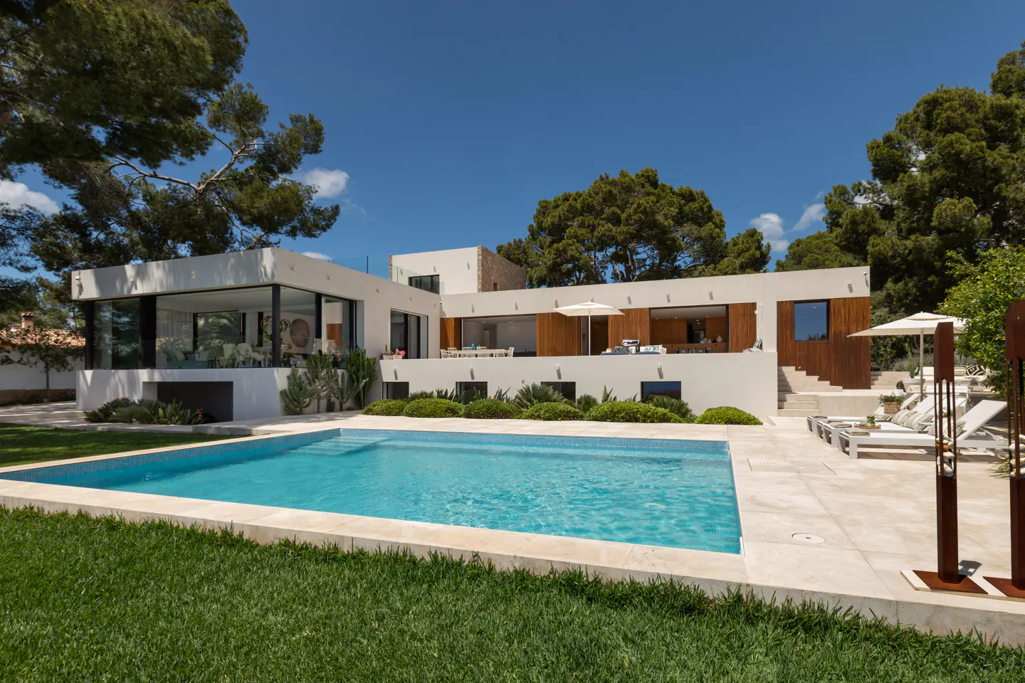 Modern white house with a blue swimming pool, green lawn, and trees under a clear blue sky.