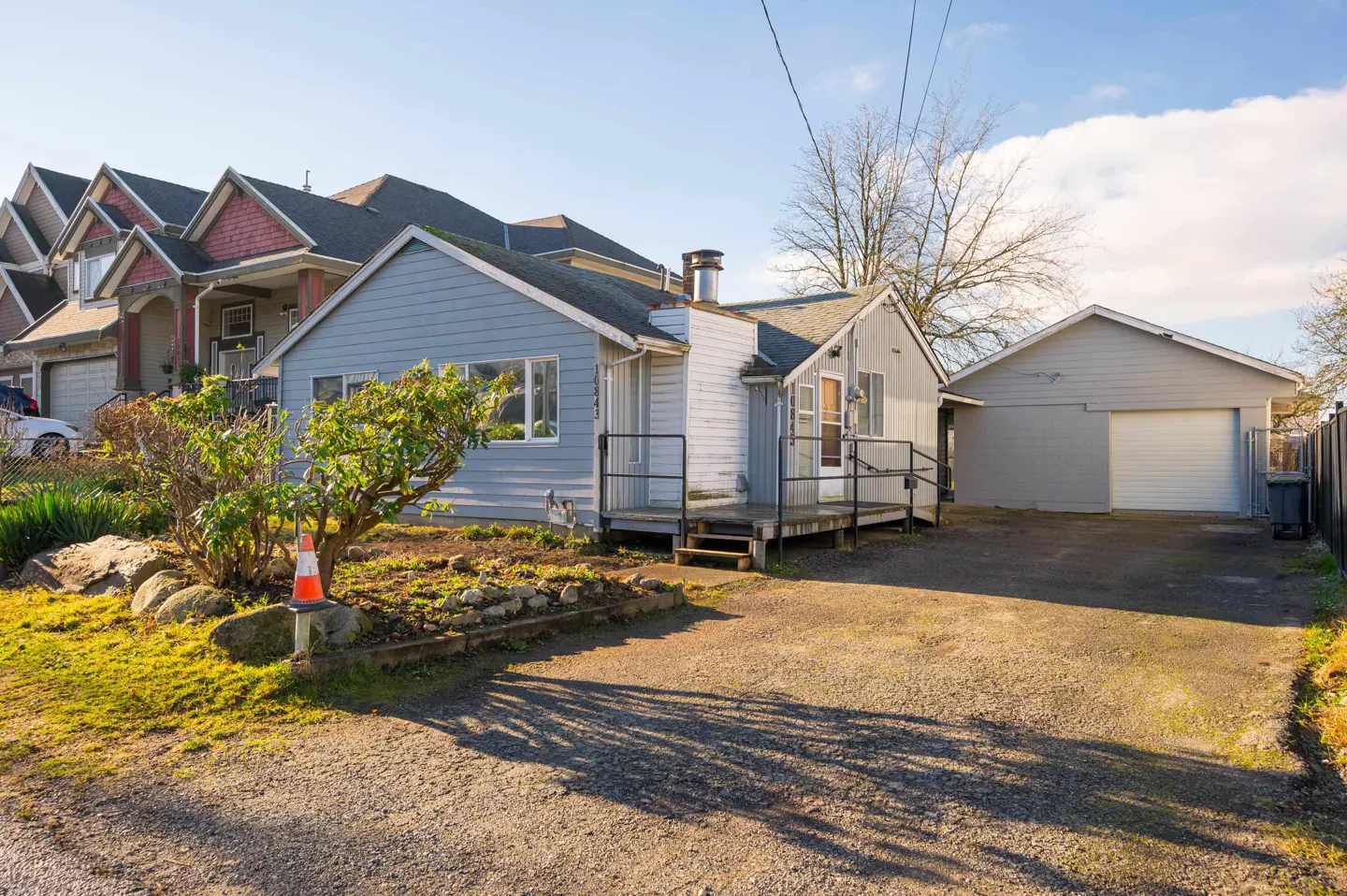 A single-story blue house with a white porch, detached garage, and gravel driveway on a sunny day.