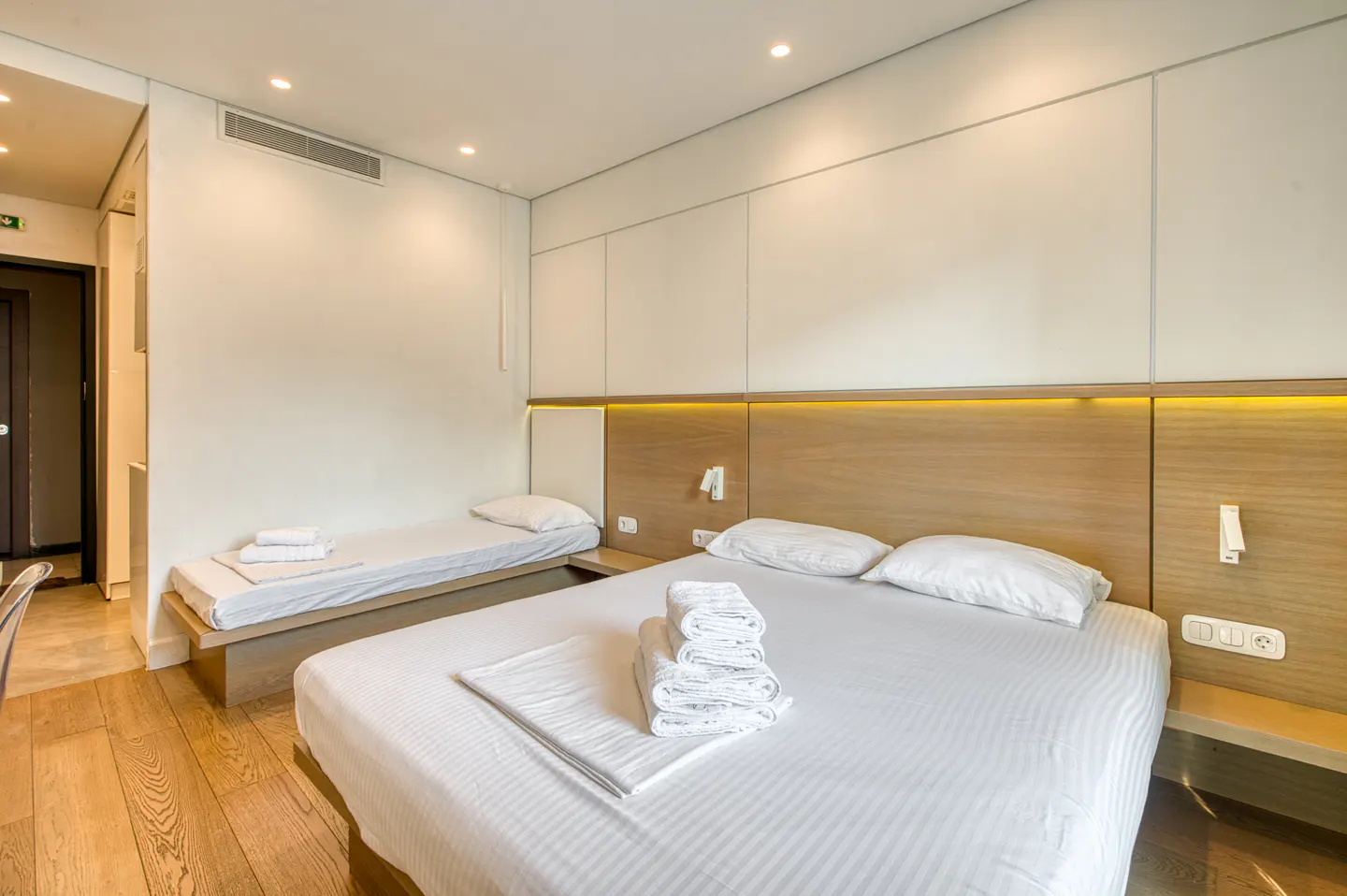 Hotel room with a queen bed and a twin bed. White walls and bedding, wood floors and headboard with yellow accent lighting.