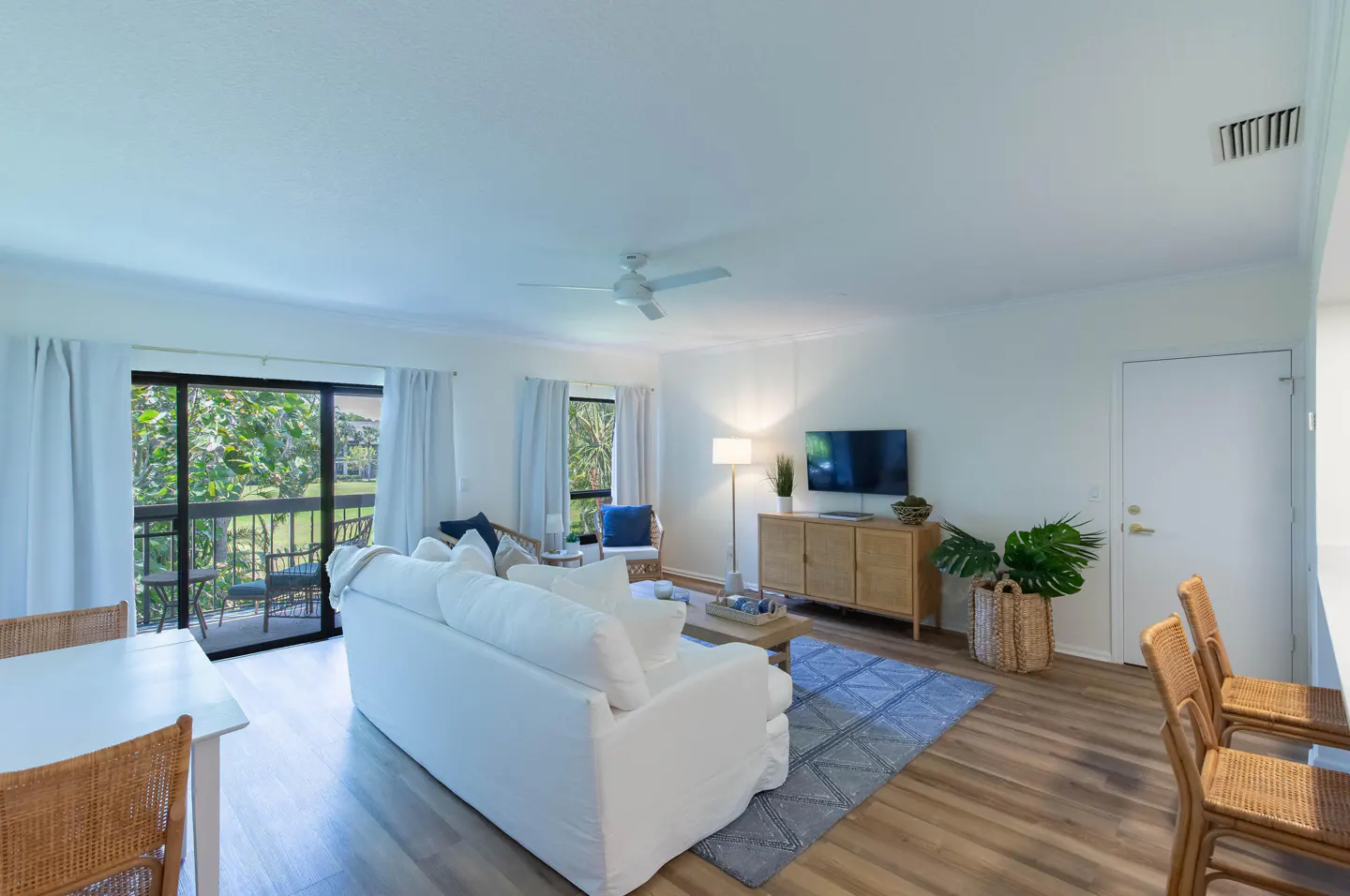 Bright living room with white sofa, wood floors, and blue rug. Sliding glass doors lead to a balcony with green foliage.