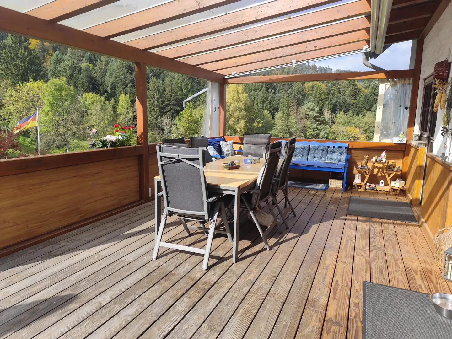 Outdoor patio with wooden deck, table, chairs, and benches. Forest and German flag visible in the background.