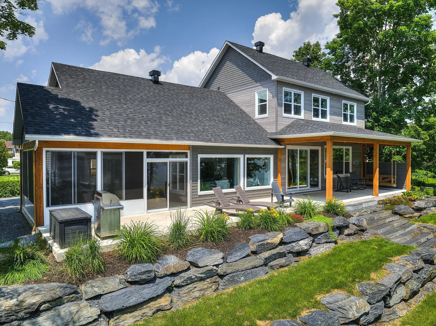 Exterior view of a gray two-story house with a screened porch, patio, and stone retaining wall.