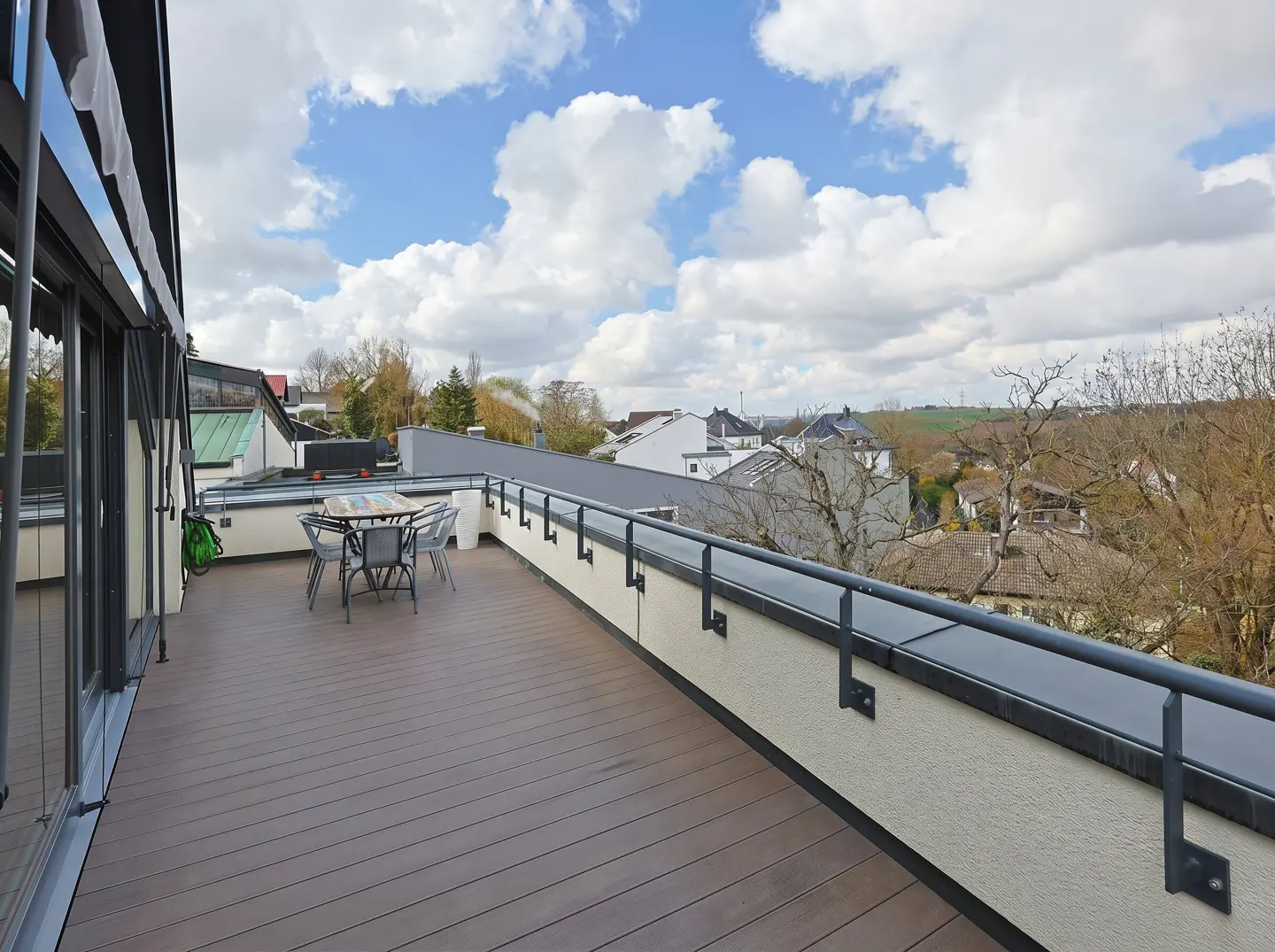 A rooftop patio with a table and chairs overlooks a neighborhood under a cloudy sky.