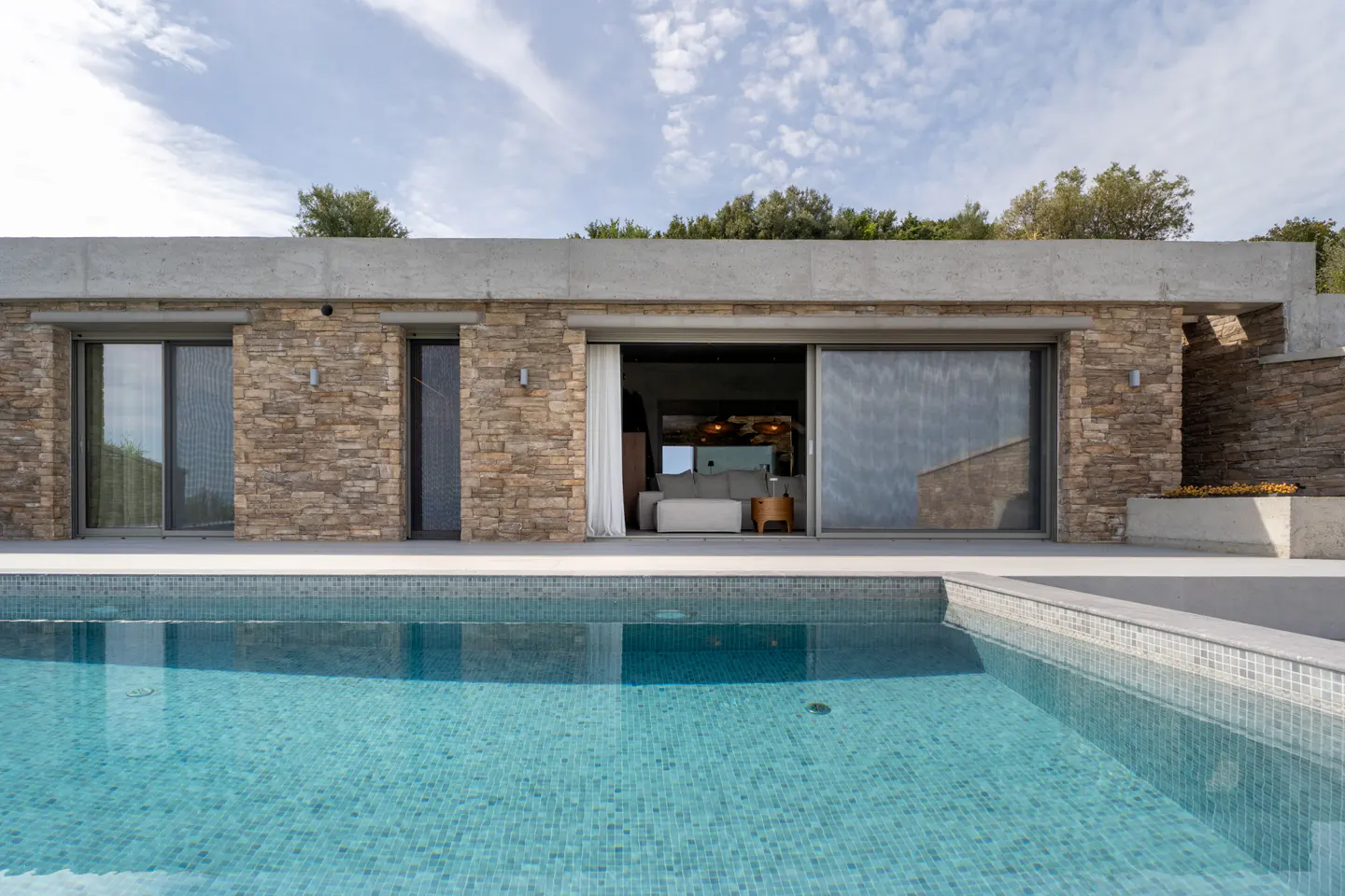 Exterior view of a stone house with a blue tiled pool in the foreground, sliding glass doors open to a modern interior.