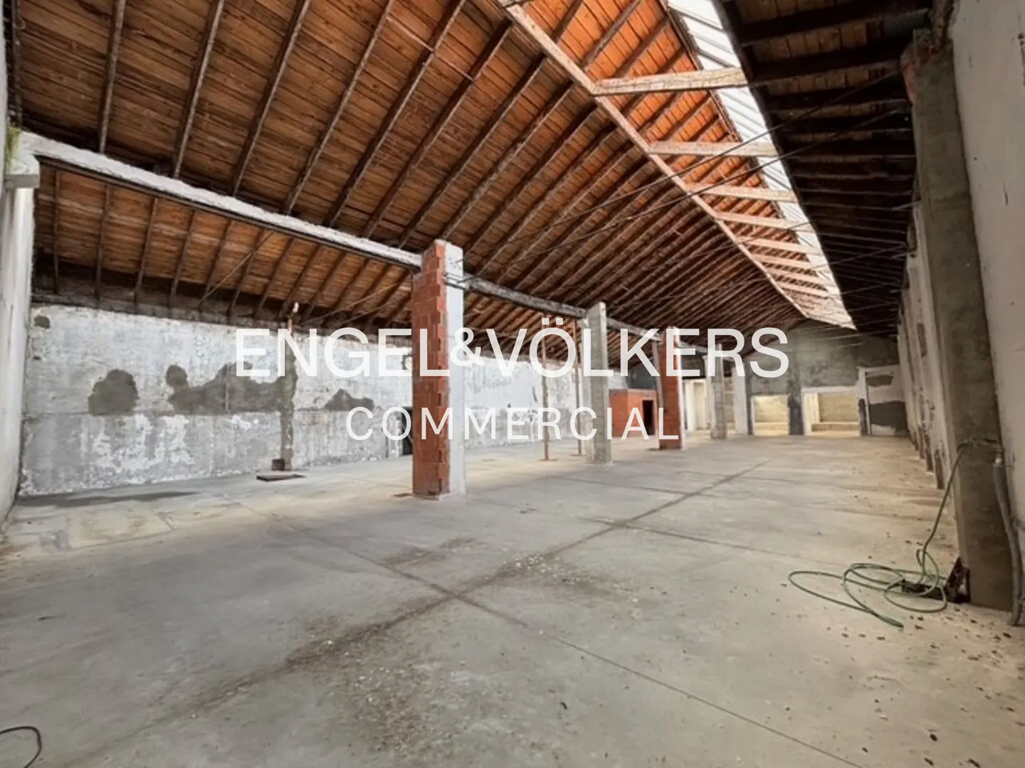Large, empty commercial space with concrete floors, brick pillars, and a high, exposed wooden beam ceiling. "Engel & Volkers Commercial" logo is superimposed.