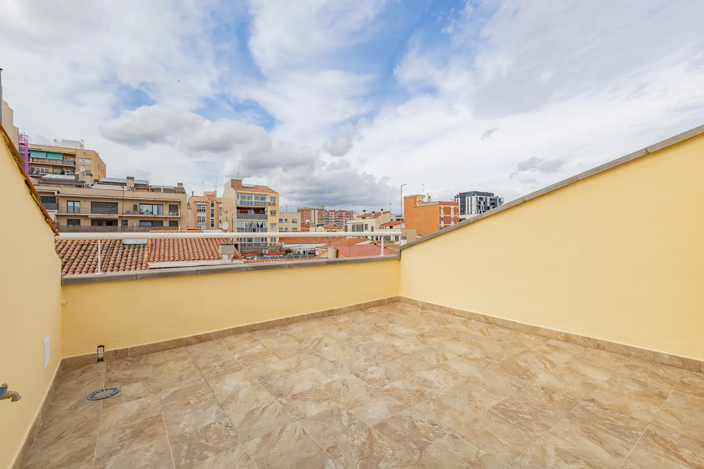 Empty rooftop terrace with beige tiled floor, yellow walls, and a white railing overlooking a city skyline under clouds.