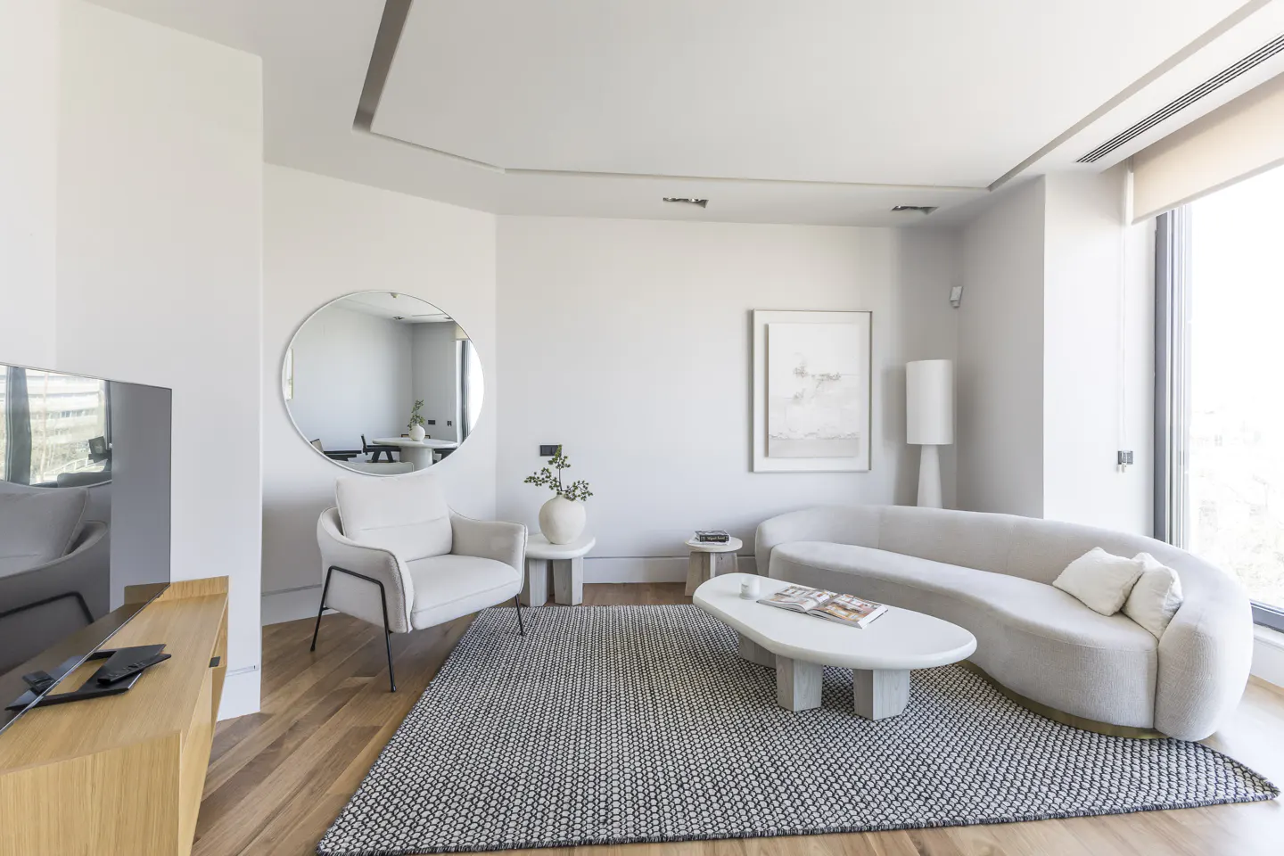Bright living room with wood floors, a gray rug, and white furniture. A round mirror reflects a dining area. A large window lets in natural light.