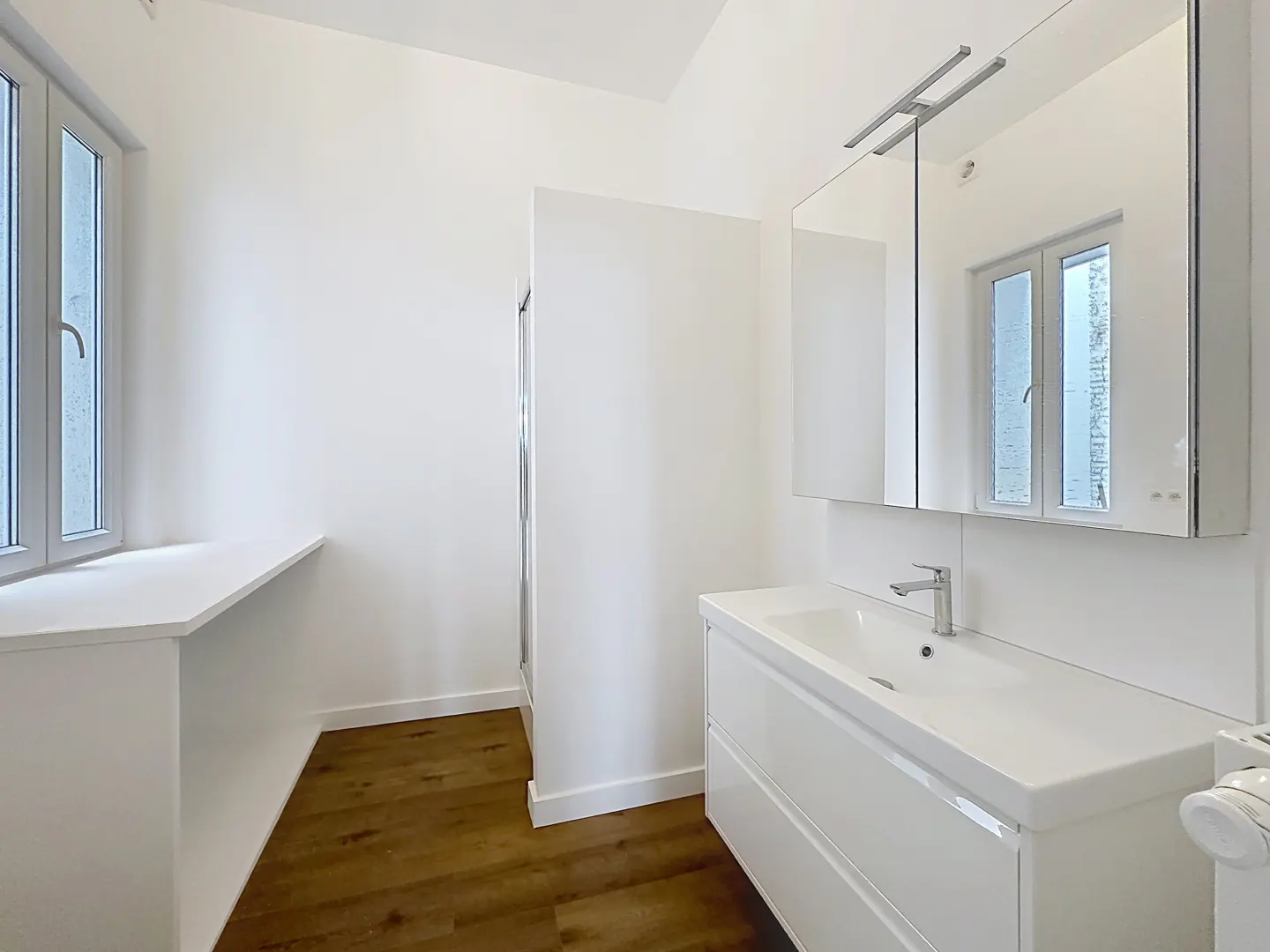 Bright, modern bathroom with white walls, vanity, and window. Wood-look flooring adds warmth. A mirrored cabinet hangs above the sink.