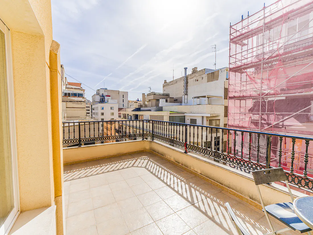 Balcony view with black iron railing, tan tile floor, and a chair and table. Buildings and scaffolding are in the background.