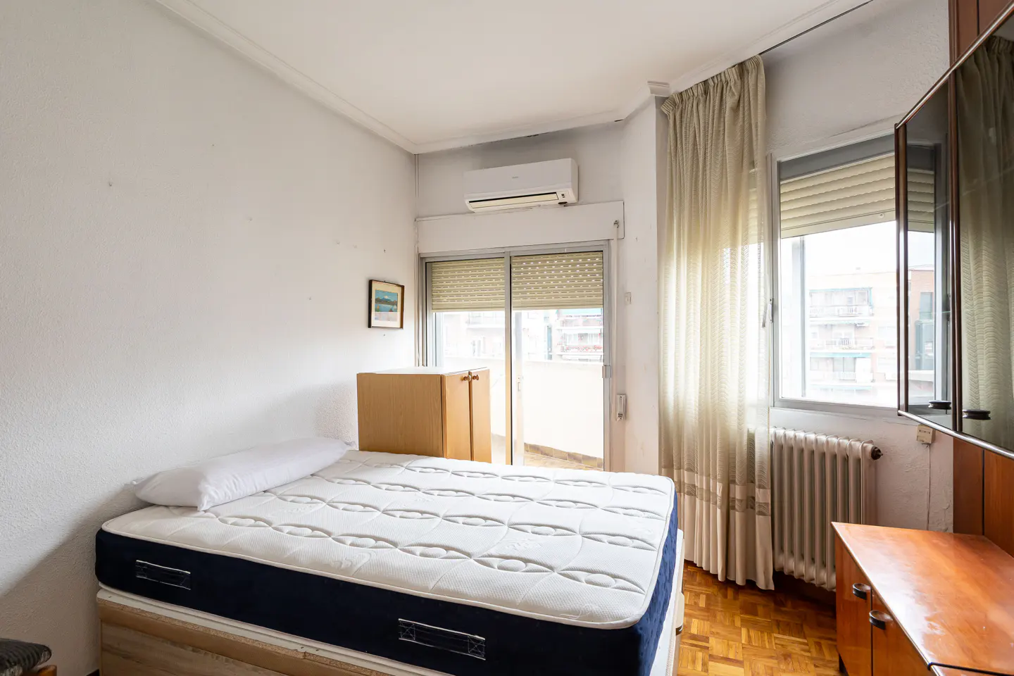 Bedroom with a bed, white walls, wood floors, and a window with sheer curtains. A cardboard box sits on the bed.