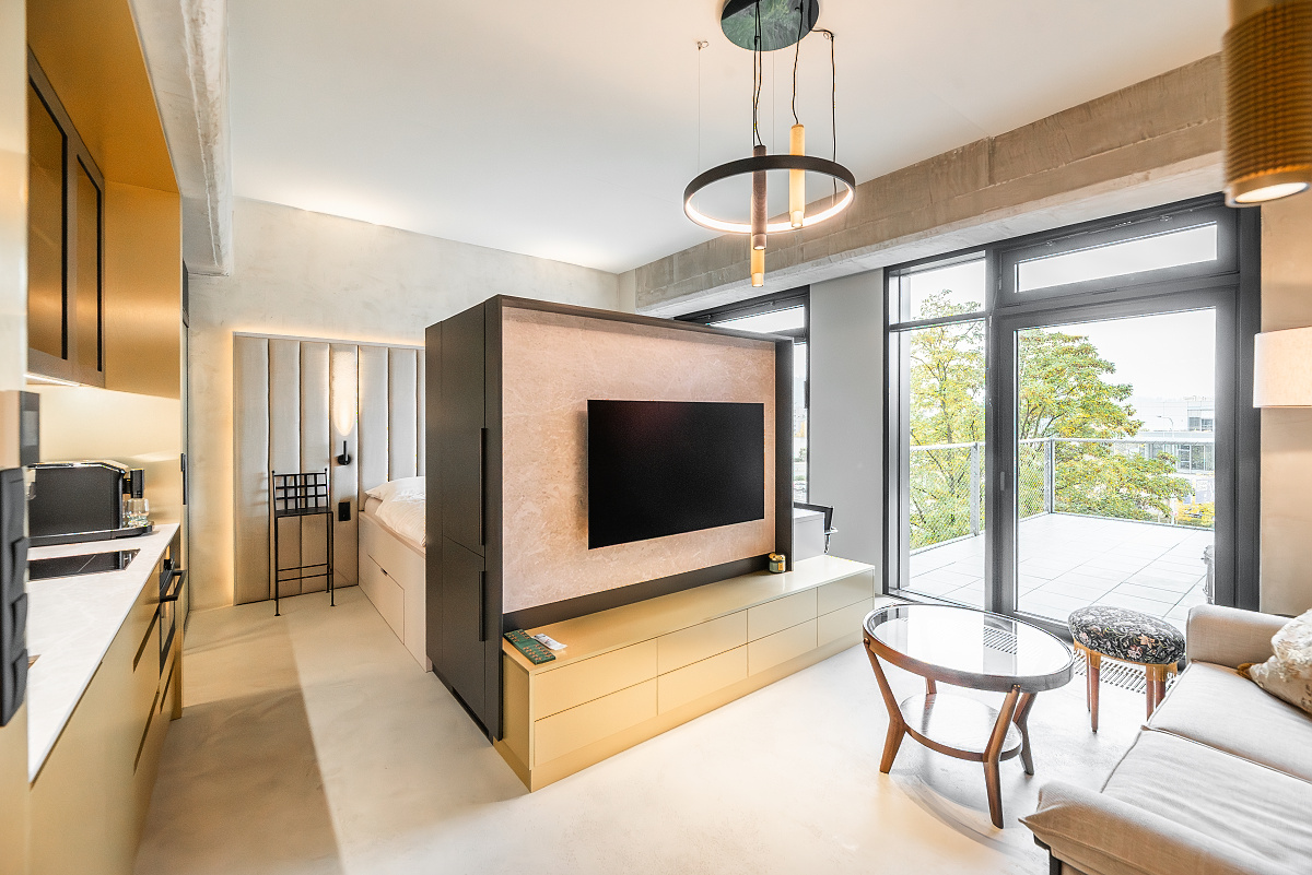 Modern studio apartment with a kitchen, a central partition wall holding a TV, a sofa, and a balcony with outdoor views. [AI-generated text]