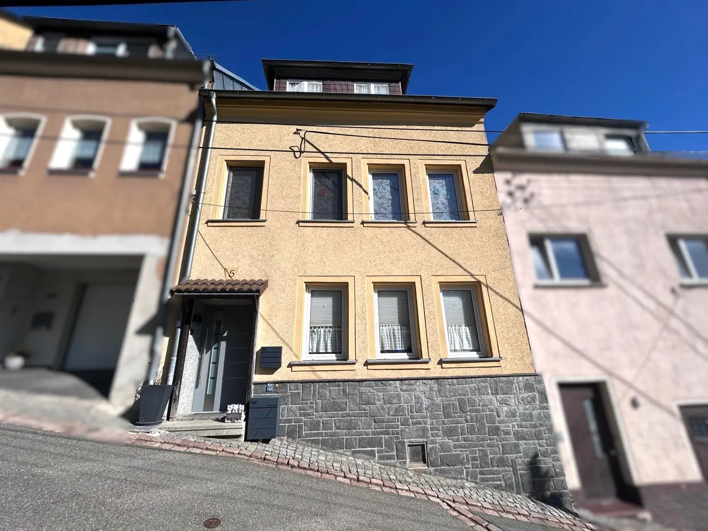 Three-story yellow house with gray stone foundation, six windows, and a gray door on a sunny day.