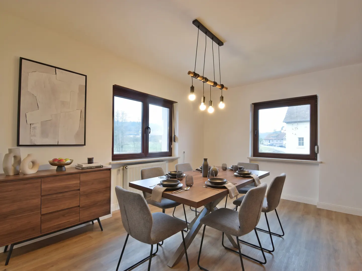 Dining room with wood table, gray chairs, and modern light fixture. A sideboard and abstract art are on the left. Windows show a rural view.