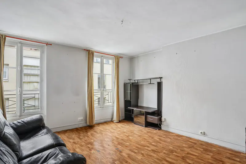 A living room with white walls, wood floors, a black leather couch, and two windows with tan curtains.