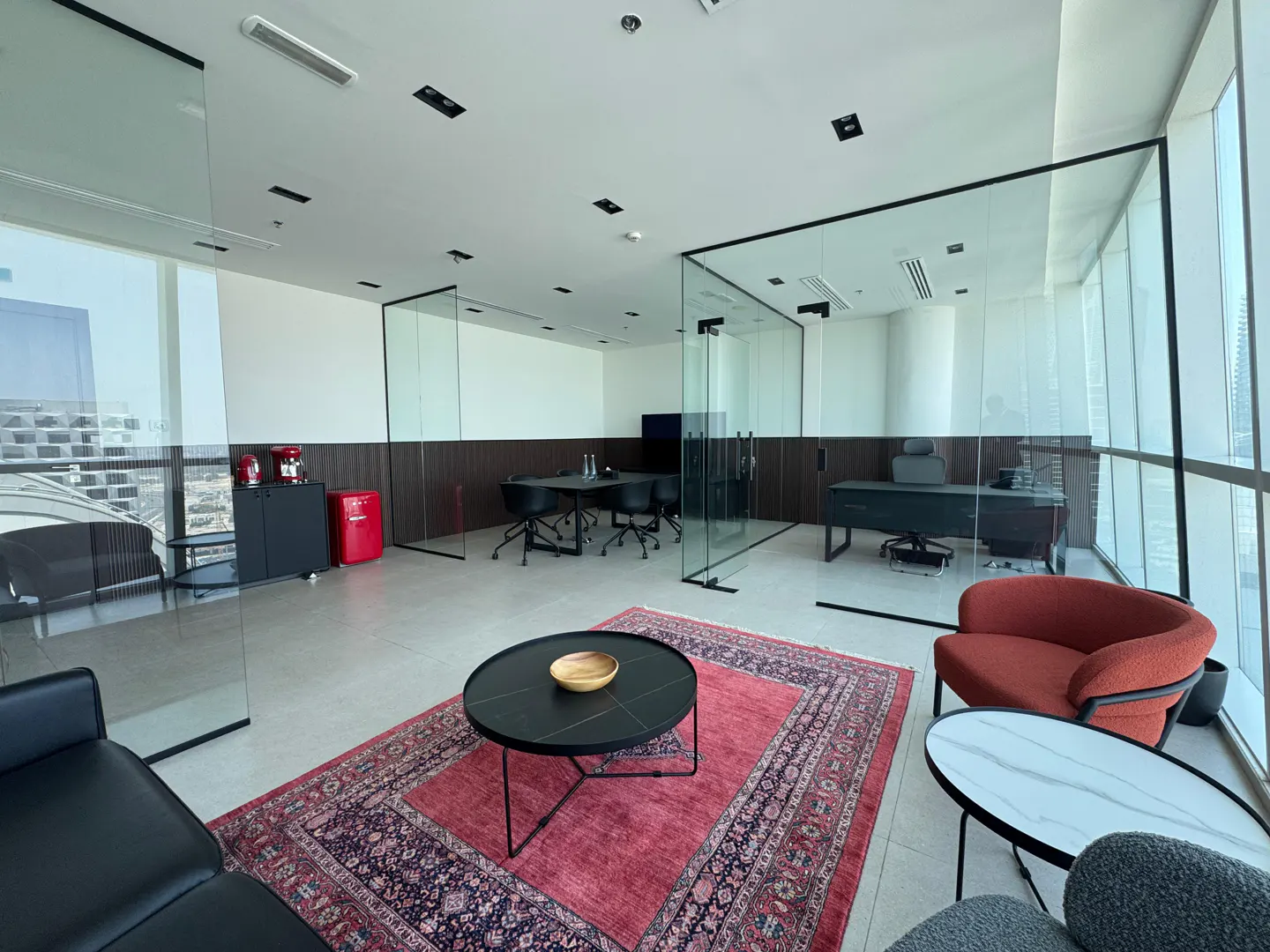 A modern office space with glass-walled rooms, a red patterned rug, and black and red furniture.