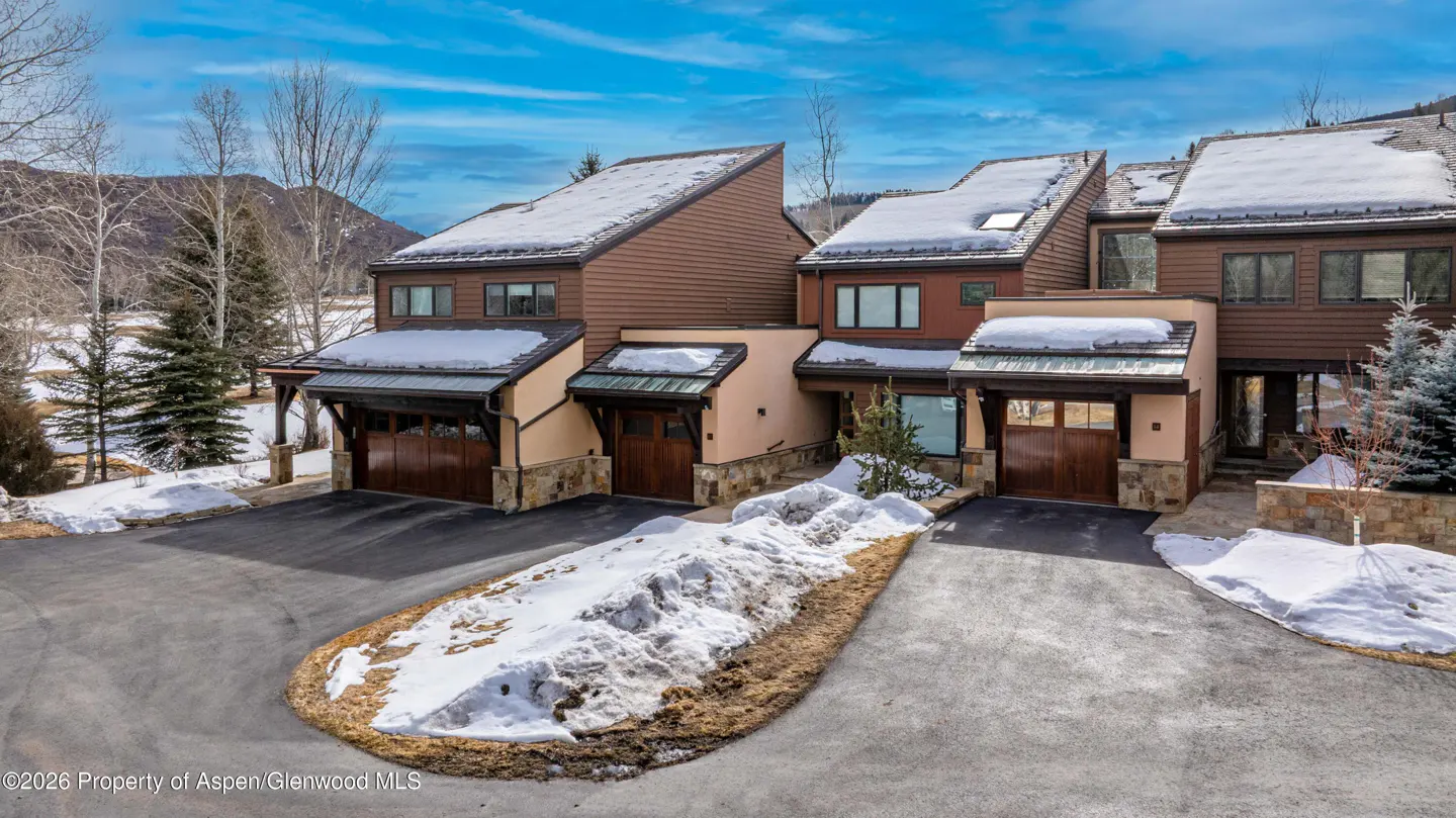 Three brown townhouses with snow-covered roofs and driveways on a sunny day.