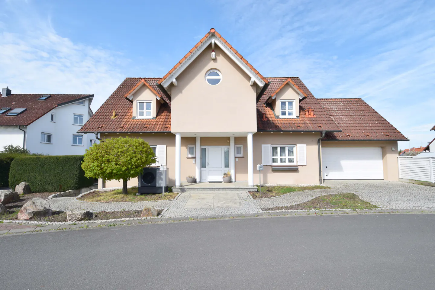 A beige two-story house with a red tile roof, white trim, and a white garage door. A green tree is in the front yard.