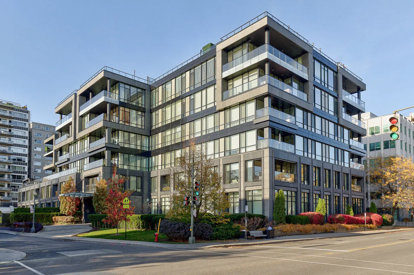 Modern gray apartment building with glass balconies and large windows, surrounded by greenery and a street view.