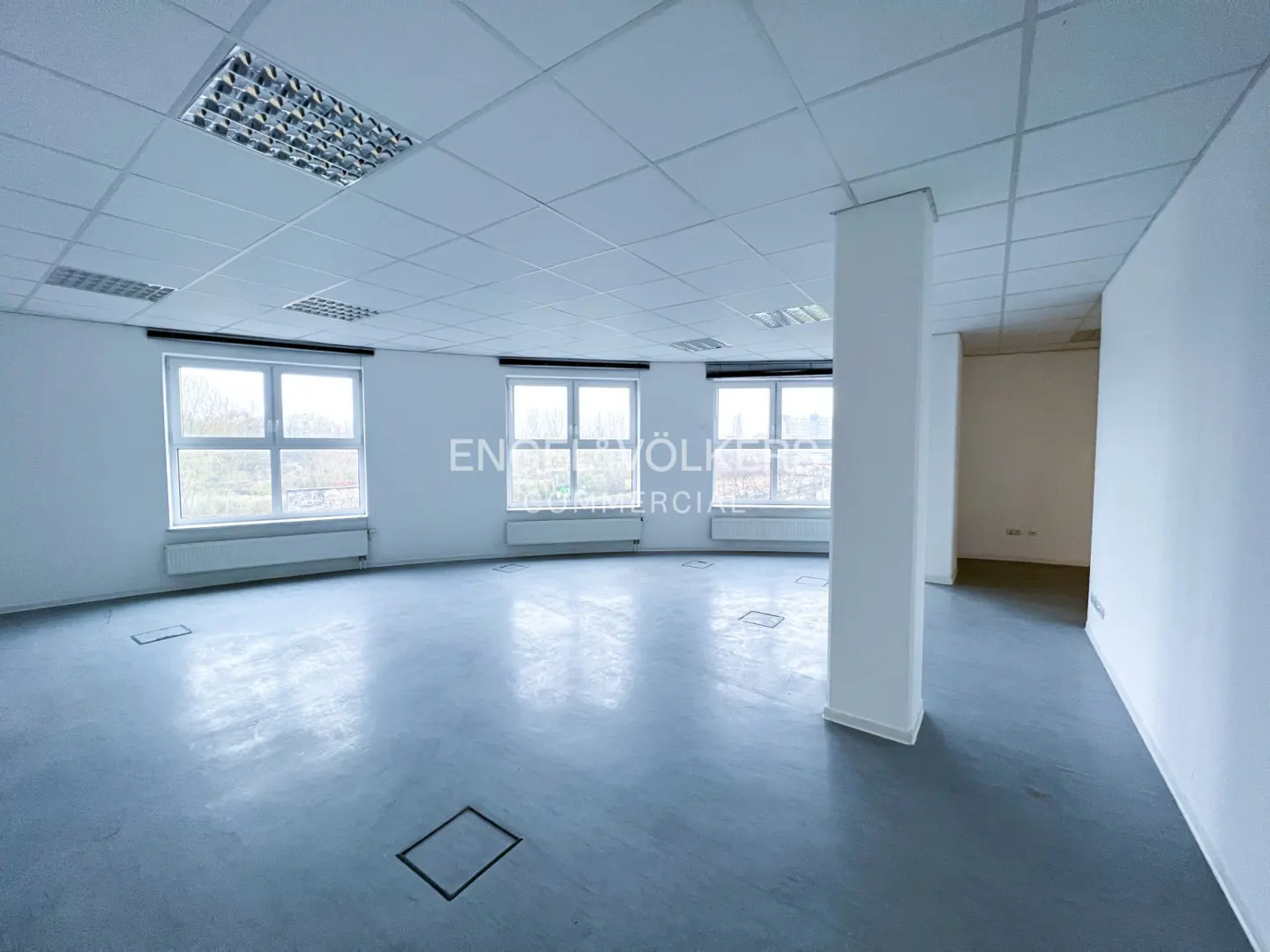 Empty, bright commercial office space with white walls, gray flooring, a drop ceiling, and three large windows.