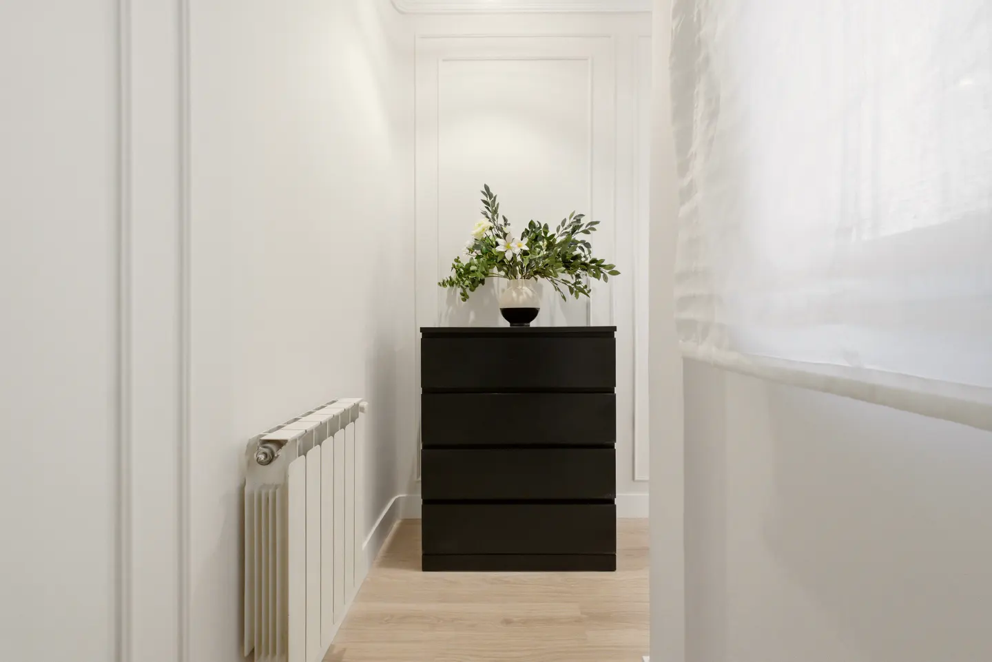 Hallway with a black four-drawer dresser topped with a floral arrangement. A white radiator is on the left, and a sheer curtain is on the right.